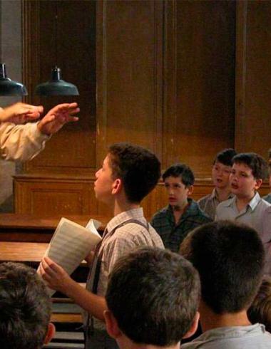 El profesor Clément Mathieu dirige un coro de niños en un salón de clases mientras varios alumnos cantan y observan atentamente durante un ensayo en una escena de la película francesa Los Coristas (Les Choristes), dirigida por Christophe Barratier.