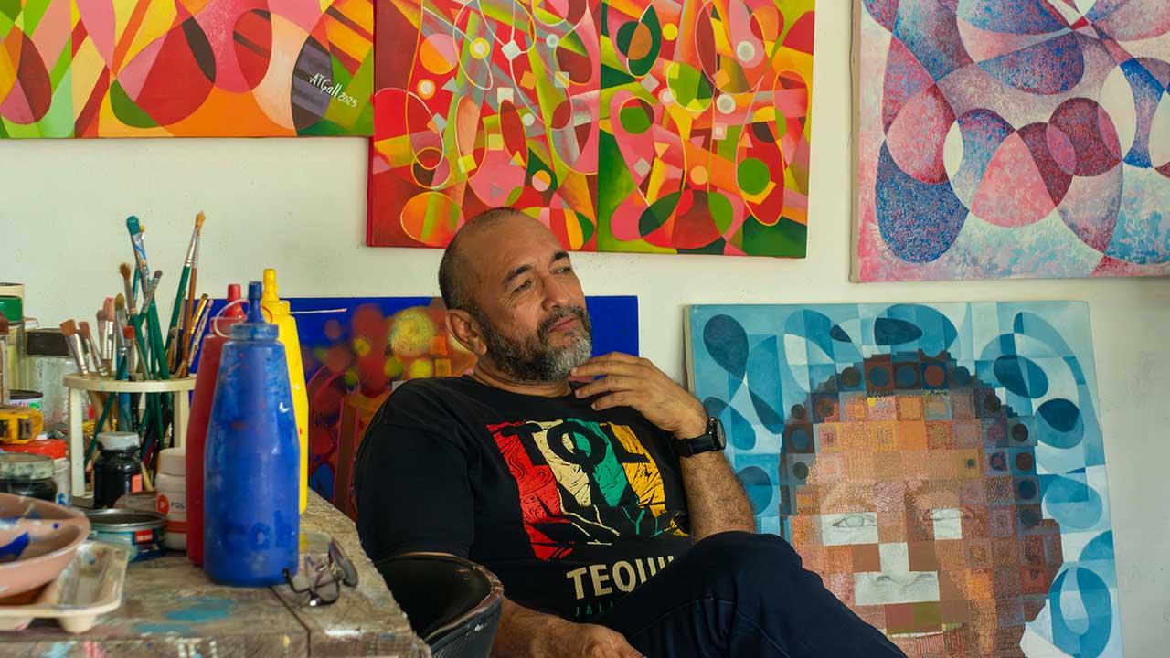 Antonio Tabáres Gallardo sentado en su taller entre pinturas abstractas coloridas, con pinceles y botellas de pintura a su lado, mirando pensativo hacia un lienzo.