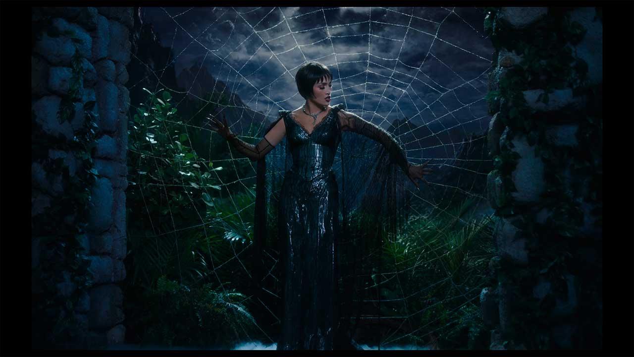 Jennifer López como La Mujer Araña en un escenario teatral, de pie frente a una telaraña gigante que refleja su silueta, vestida con un traje brillante de lentejuelas.
