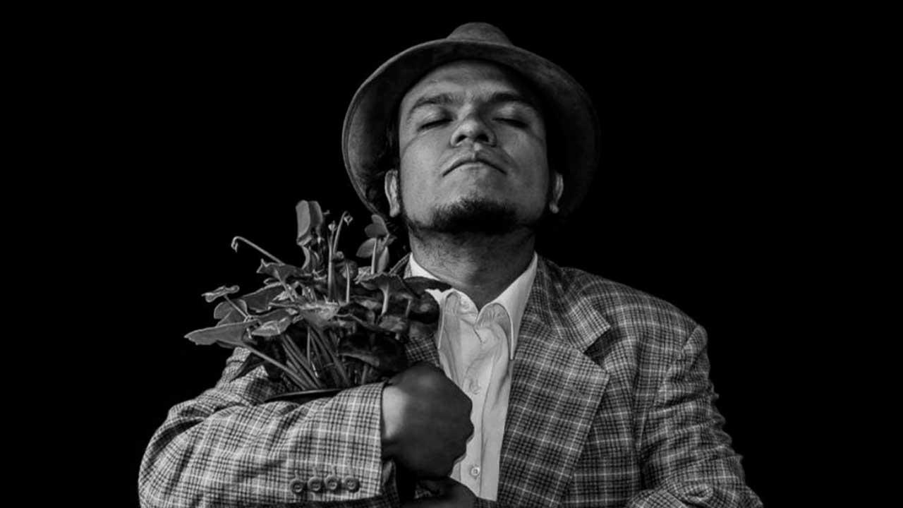 Retrato en blanco y negro del actor y director teatral Pedro Acevedo, con sombrero y saco a cuadros, sosteniendo un ramo de plantas entre sus brazos mientras mantiene los ojos cerrados en una pose introspectiva sobre un fondo negro.