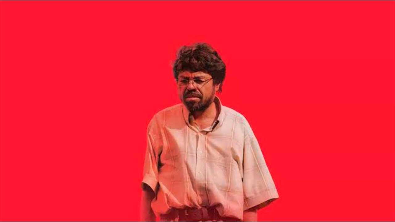 Un hombre de lentes y cabello alborotado con camisa de manga corta mirando de frente sobre un fondo rojo intenso, fotograma de la película Un Poeta de Simón Mesa Soto.