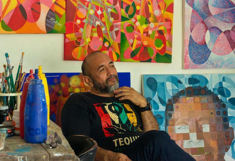 Antonio Tabáres Gallardo sentado en su taller entre pinturas abstractas coloridas, con pinceles y botellas de pintura a su lado, mirando pensativo hacia un lienzo.