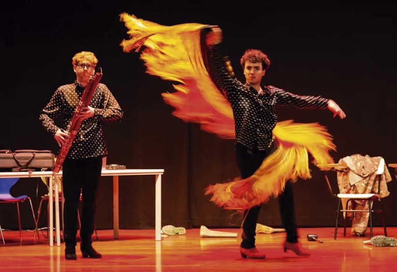 Dos artistas se presentan sobre un escenario; uno interpreta un instrumento de viento de madera mientras el otro realiza un movimiento de danza con una larga pieza de tela amarilla y naranja.