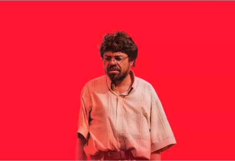 Un hombre de lentes y cabello alborotado con camisa de manga corta mirando de frente sobre un fondo rojo intenso, fotograma de la película Un Poeta de Simón Mesa Soto.