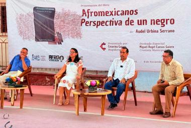 Audel Urbina Serrano presenta su libro "Afromexicanos, perspectiva de un negro" en Coyuca de Benítez, acompañado por la regidora Cesarina Quinto, el cronista Miguel Ángel Cuevas Quinto y otros invitados, frente al banner oficial del evento.