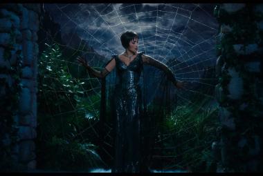 Jennifer López como La Mujer Araña en un escenario teatral, de pie frente a una telaraña gigante que refleja su silueta, vestida con un traje brillante de lentejuelas.