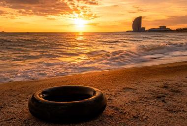 Una llanta de cámara de camión descansa sobre la arena de una playa al atardecer. El sol ilumina el horizonte con tonos dorados mientras el mar se extiende hacia una ciudad costera al fondo, evocando un paisaje de memoria, viaje y contemplación.