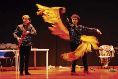 Dos artistas se presentan sobre un escenario; uno interpreta un instrumento de viento de madera mientras el otro realiza un movimiento de danza con una larga pieza de tela amarilla y naranja.