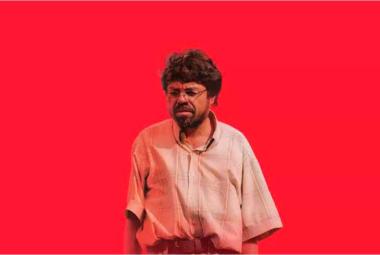 Un hombre de lentes y cabello alborotado con camisa de manga corta mirando de frente sobre un fondo rojo intenso, fotograma de la película Un Poeta de Simón Mesa Soto.