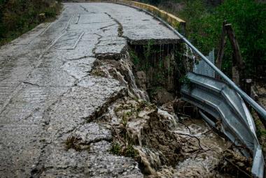 Fotografía de una carretera de concreto rural gravemente colapsada en un acantilado bajo una fuerte lluvia. Una parte significativa del pavimento se ha desmoronado. El agua de lluvia fluye activamente por las grietas y las caras expuestas de la tierra y la roca, y cae sobre un culvert de metal oxidado al pie del colapso. Árboles verdes se alinean en la colina bajo el clima lluvioso.