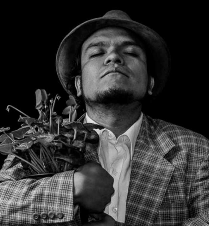 Retrato en blanco y negro del actor y director teatral Pedro Acevedo, con sombrero y saco a cuadros, sosteniendo un ramo de plantas entre sus brazos mientras mantiene los ojos cerrados en una pose introspectiva sobre un fondo negro.