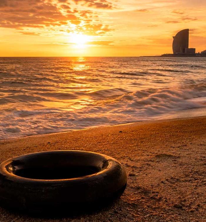 Una llanta de cámara de camión descansa sobre la arena de una playa al atardecer. El sol ilumina el horizonte con tonos dorados mientras el mar se extiende hacia una ciudad costera al fondo, evocando un paisaje de memoria, viaje y contemplación.