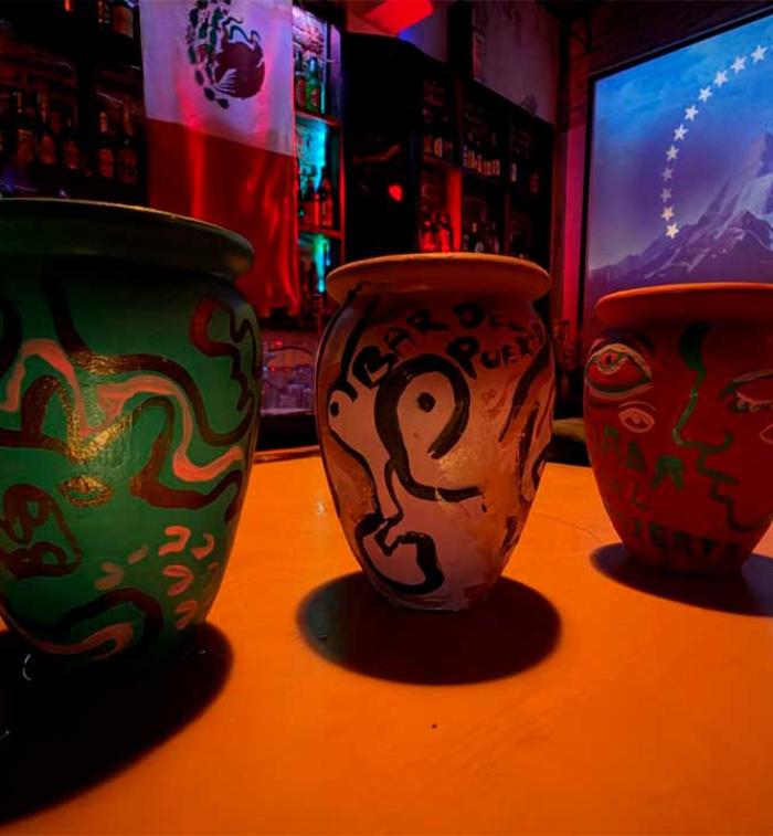 Tres cantaritos de barro pintados a mano sobre la barra del Bar del Puerto, iluminados con luces de colores y con una proyección al fondo.